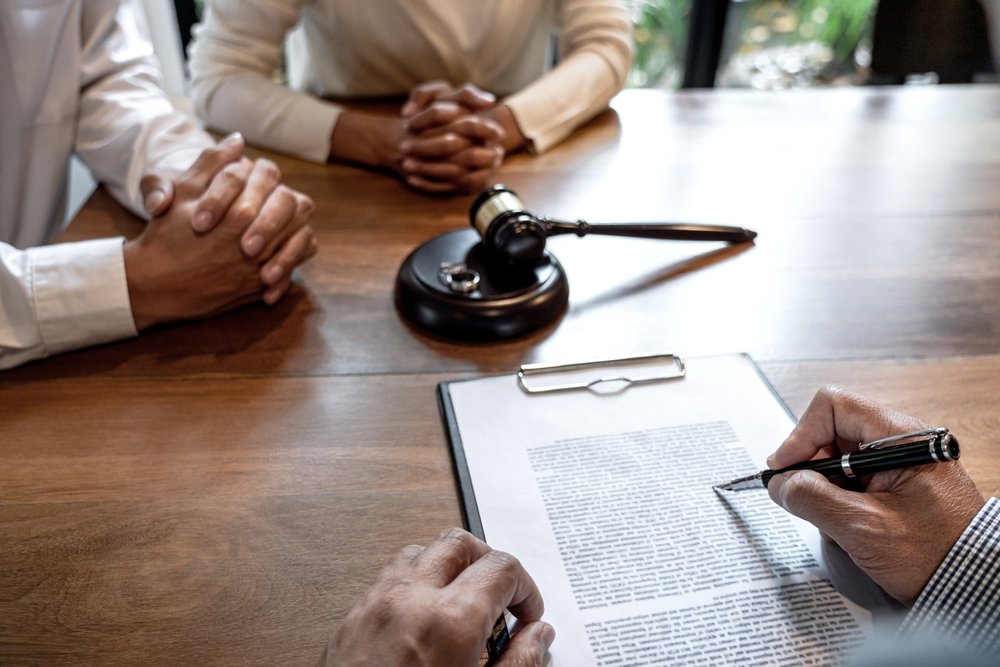 Divorcing couple sitting across from attorney signing paperwork with gavel and wedding rings on desk