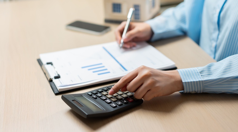 Person in a striped blue shirt using a calculator and taking notes on a clipboard with financial charts, with a small model house on the desk