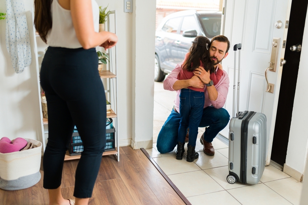 Father kneeling to hug his young daughter at the front door beside a rolling suitcase while her mother looks on